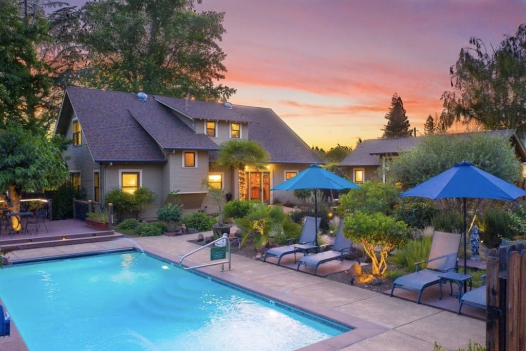 A serene backyard oasis with a sparkling pool, surrounded by lush greenery and comfortable lounge chairs under blue umbrellas, as the sun sets behind a charming two-story house in Healdsburg, California.