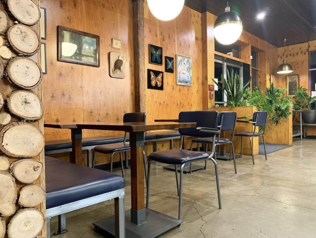 Plank Coffee A cozy cafe with wooden walls adorned with framed art, featuring tables, chairs, and a bench with blue cushions, illuminated by hanging pendant lights in Healdsburg, California.
