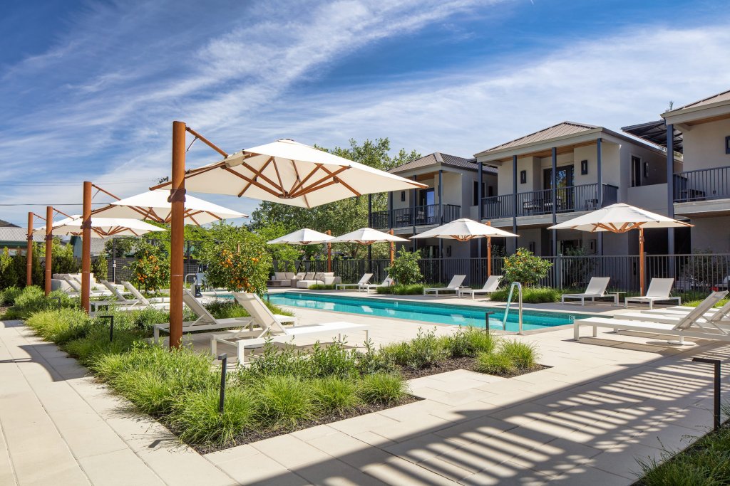 A serene outdoor pool area with multiple white umbrellas and lounge chairs, surrounded by lush greenery and modern apartment buildings in Healdsburg, California.