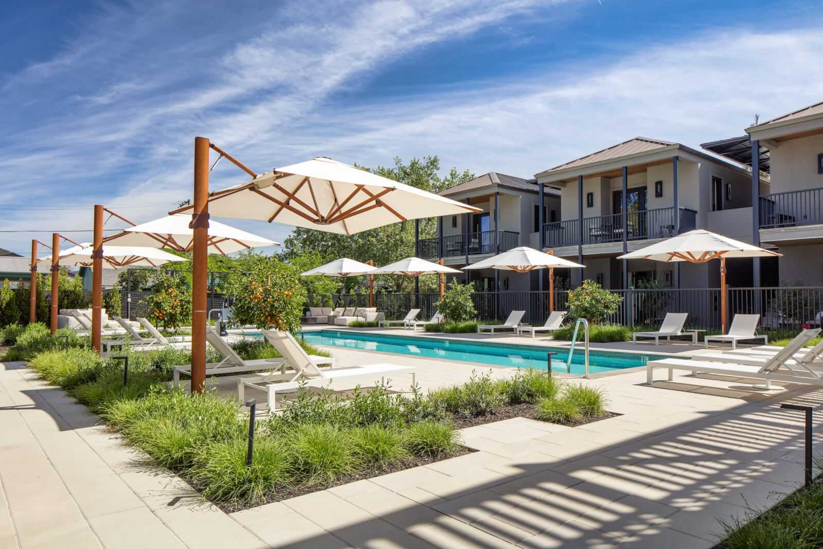 Duchamp A serene outdoor pool area with multiple white umbrellas and lounge chairs, surrounded by lush greenery and modern apartment buildings in Healdsburg, California.