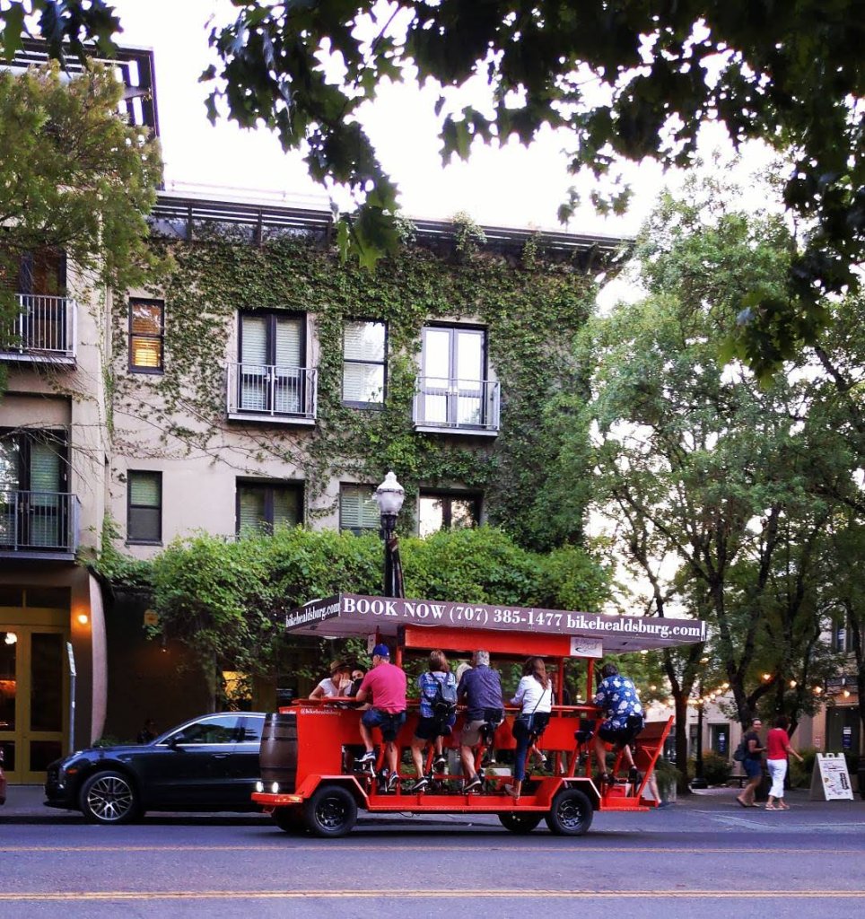 A red party bike with passengers is parked on a street next to a building covered in ivy in Healdsburg, California.