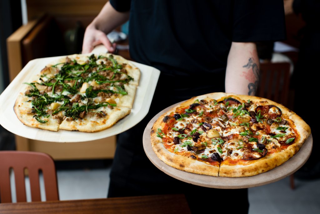 A person holds two different pizzas: one with greens and sausage, the other with olives and cheese in Healdsburg, California.