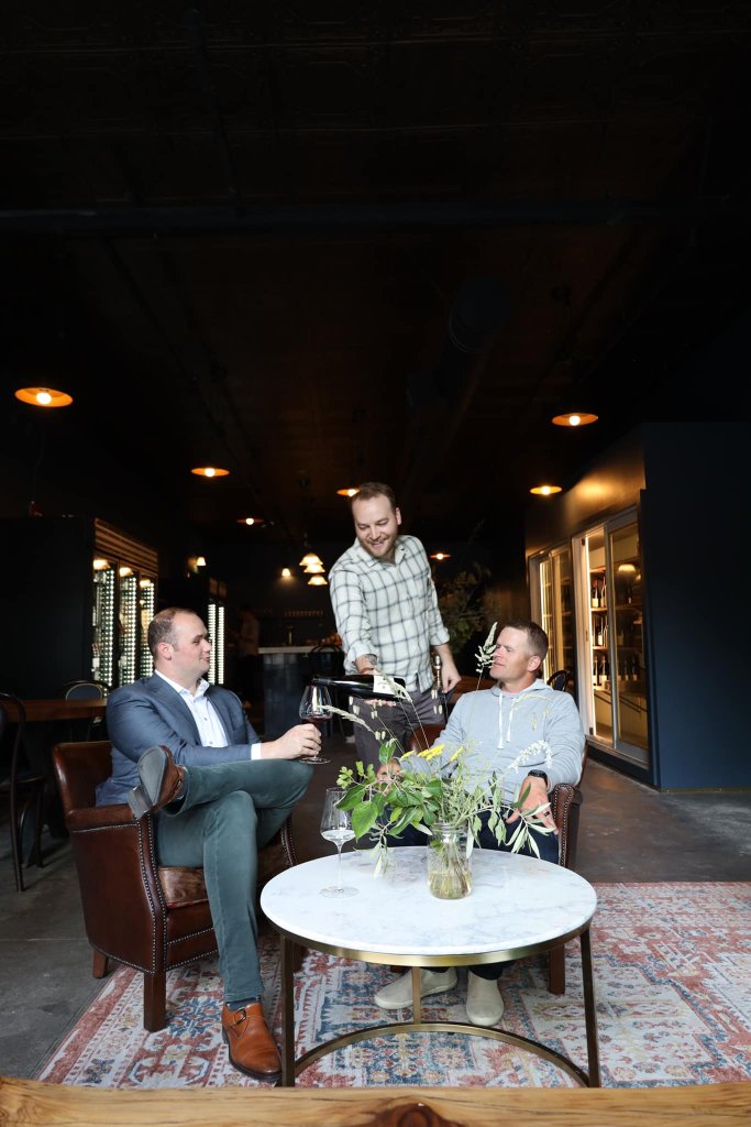 Three men in a dimly lit room, one serving wine to the others seated in leather chairs in Healdsburg, California.