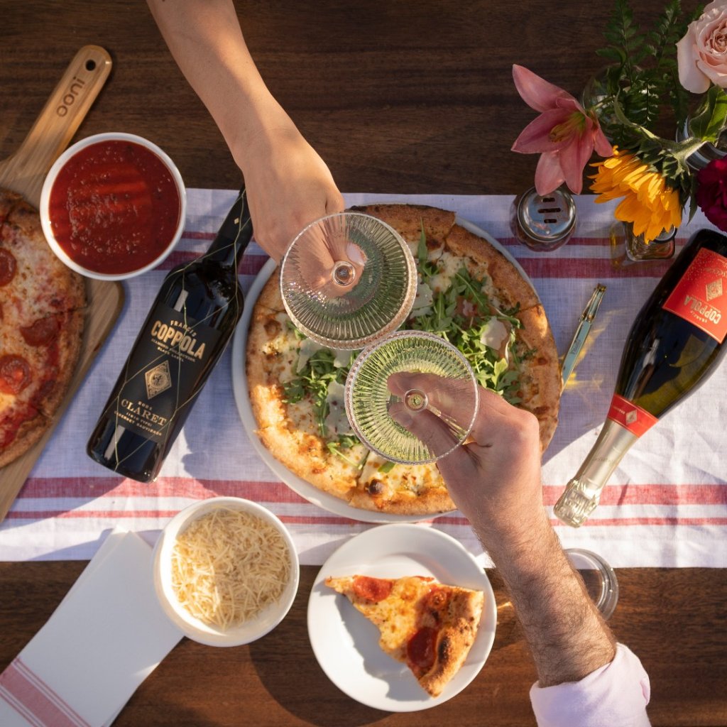 Two people clinking glasses of sparkling wine over a table set with pizza, cheese, and wine bottles in Healdsburg, California.