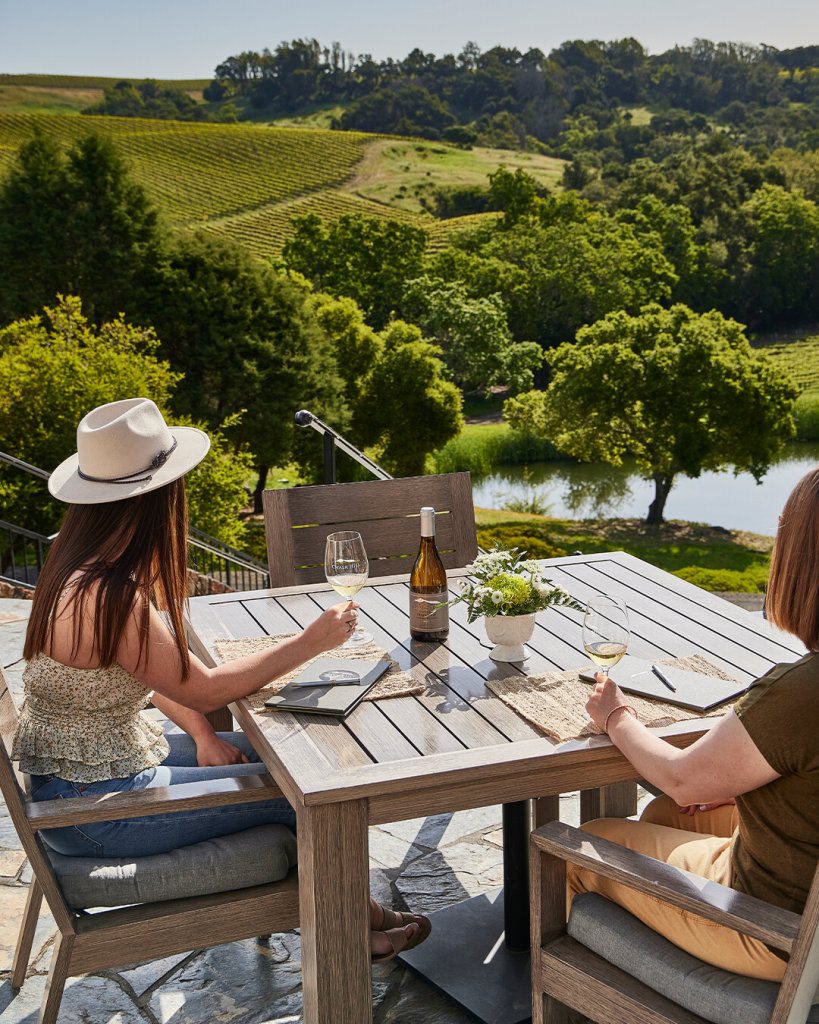 Two women enjoy wine and conversation at a table overlooking a vineyard and river in Healdsburg, California.