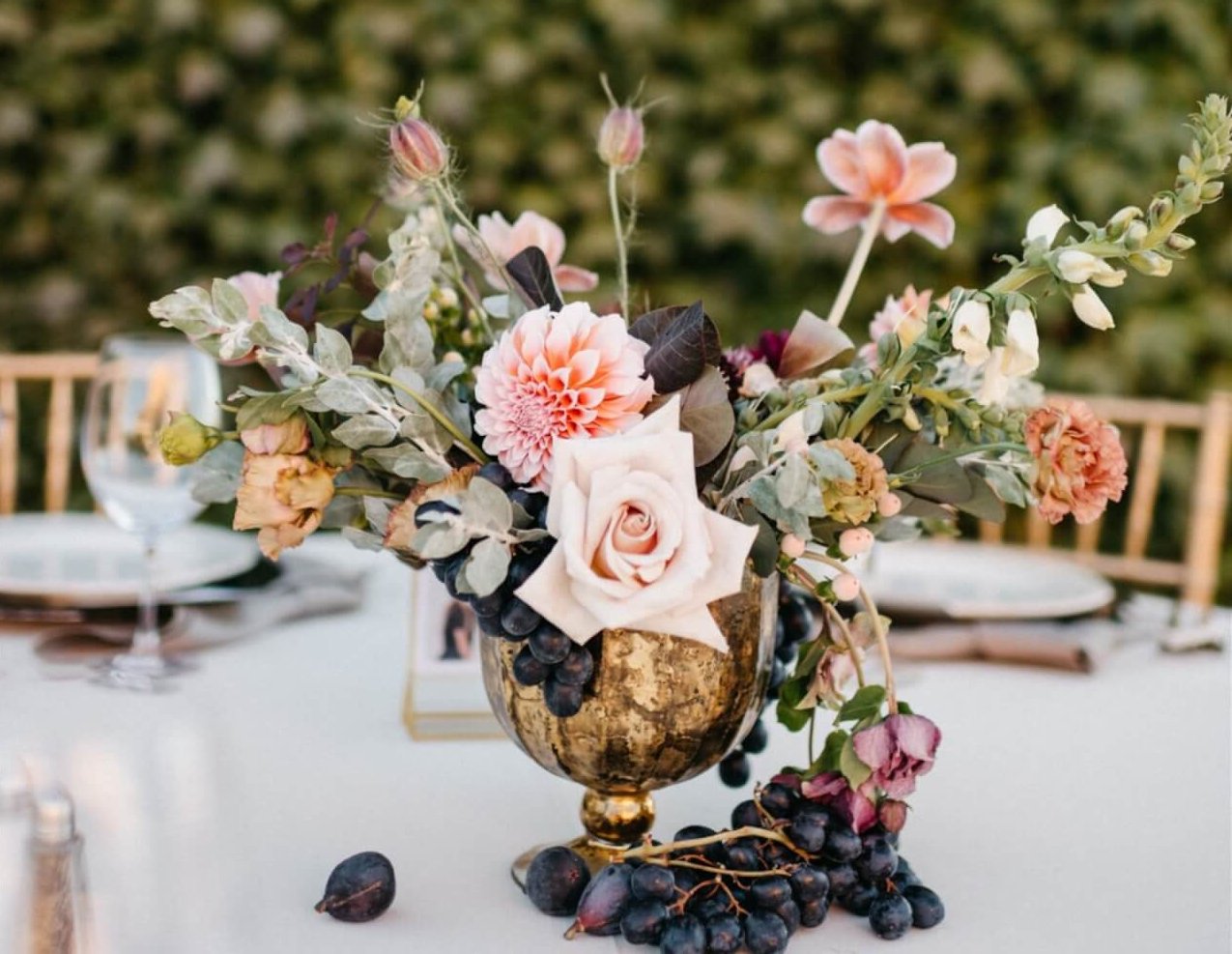 Aimee Lomeli Designs A golden vase filled with a bouquet of pink roses, dahlias, and grapes on a table in Healdsburg, California.