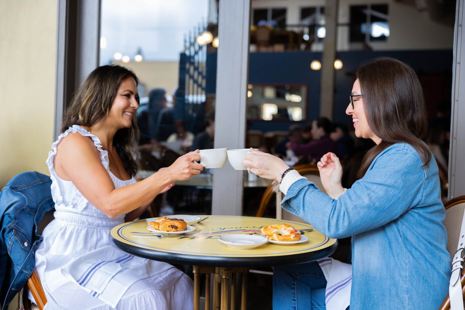 Panadería francesa Costeaux Two women clink coffee cups at an outdoor cafe table with pastries in Healdsburg, California.