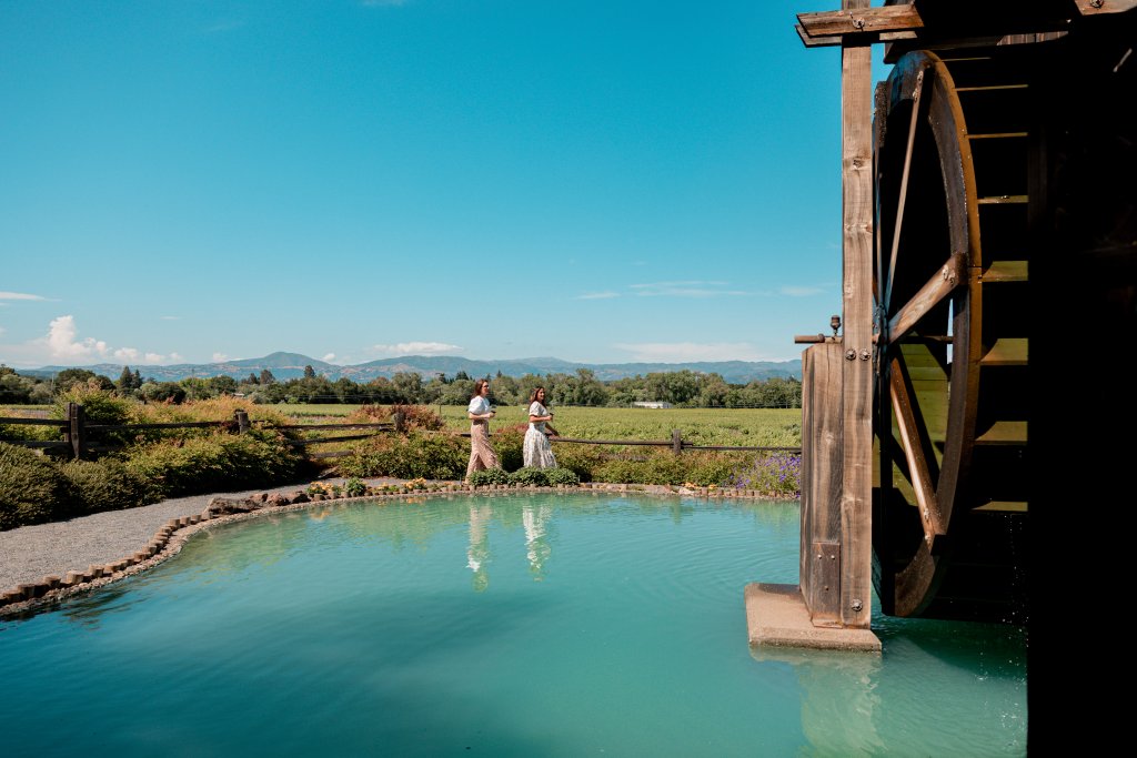 Two women stand by a serene pond with a wooden waterwheel, surrounded by lush greenery in Healdsburg, California.