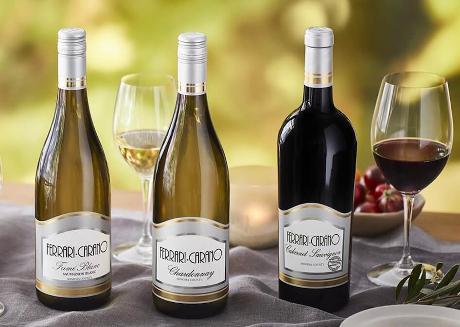 Three elegant bottles of Ferrari-Carano wine, including Fumé Blanc, Chardonnay, and Cabernet Sauvignon, are displayed alongside filled wine glasses on a table draped with a gray cloth, with a backdrop of lush greenery in Healdsburg, California.