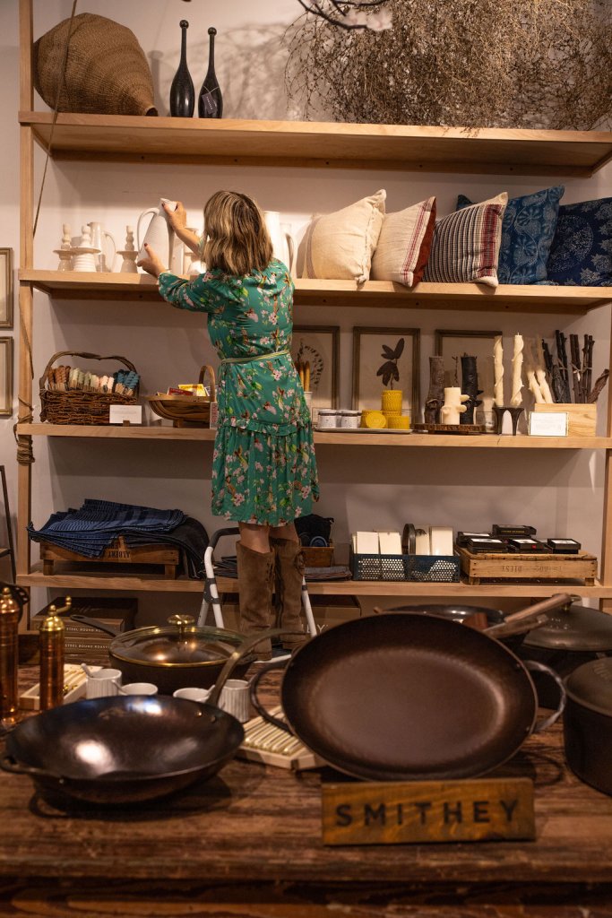 Woman in green dress arranges items on shelves in a rustic kitchen store in Healdsburg, California.