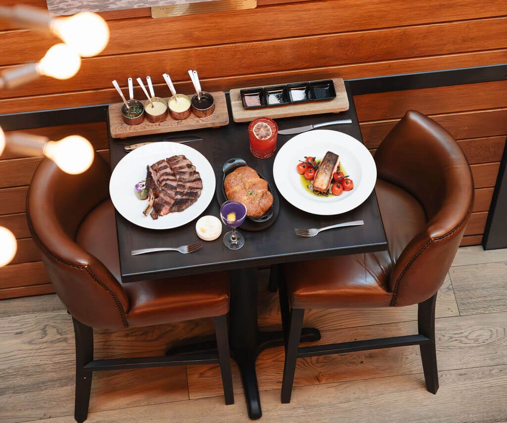 Goodnight's Prime Steak + Spirits A rustic restaurant table with two leather chairs, plates of steak and fish, and a cowboy-themed painting above in Healdsburg, California.