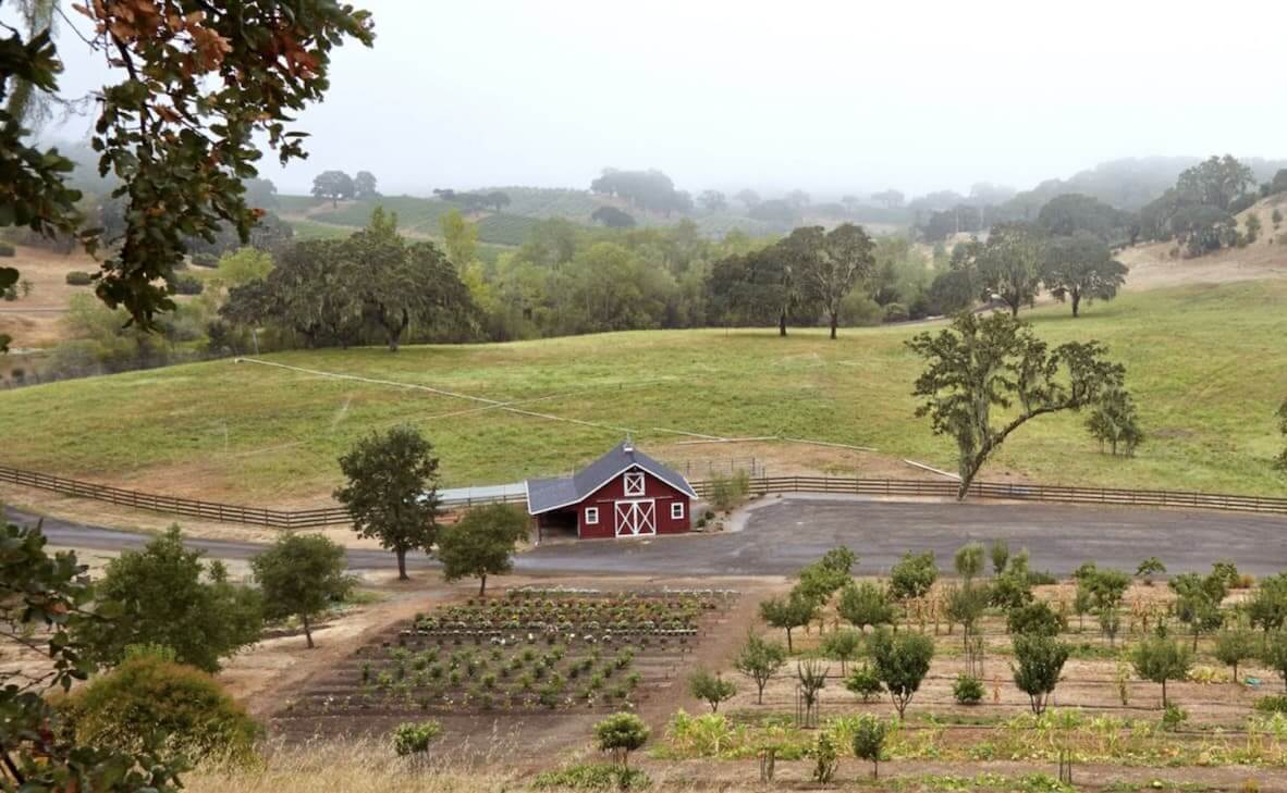 Viñedo y bodega Jordan A picturesque red barn with a white door sits in the middle of a lush green field, surrounded by rolling hills and a winding dirt road in Healdsburg, California.