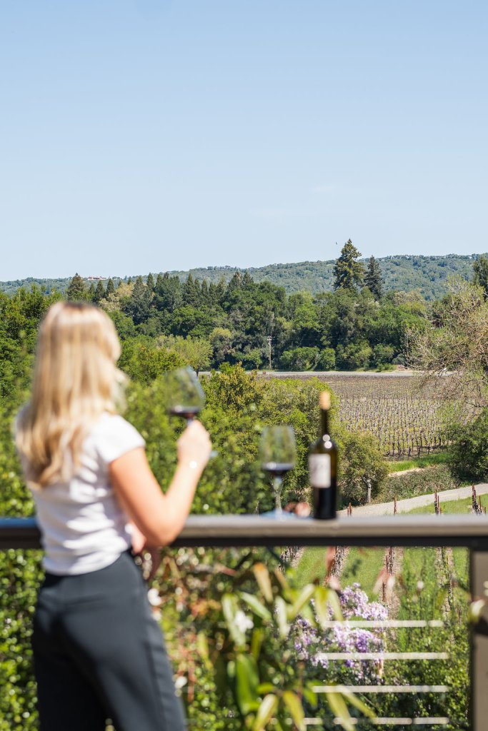 A woman with blonde hair enjoys a glass of wine on a balcony overlooking a vineyard in Healdsburg, California.