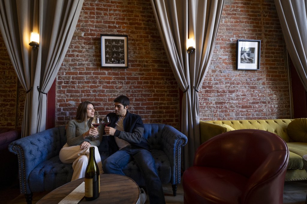 A couple enjoys wine on a blue couch in a cozy room with a brick wall and curtains in Healdsburg, California.