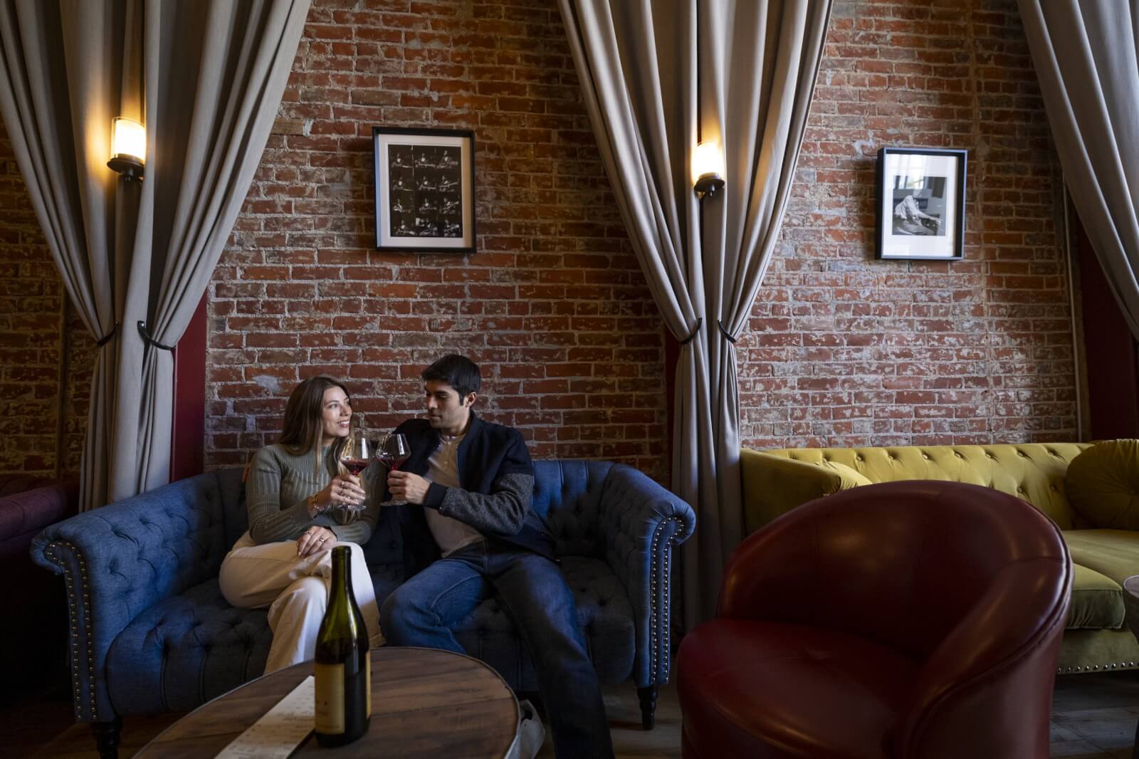 Furthermore Wines A couple enjoys wine on a blue couch in a cozy room with a brick wall and curtains in Healdsburg, California.