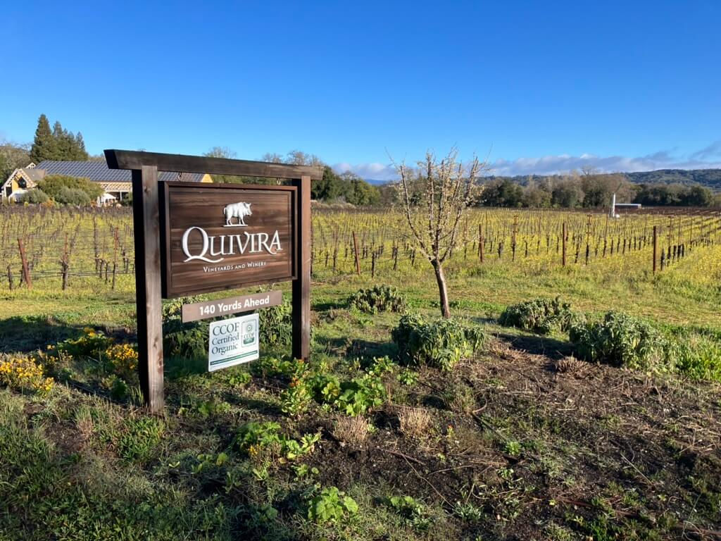 A wooden sign for the Quivira vineyard stands in a grassy field with rows of grapevines in the background in Healdsburg, California.