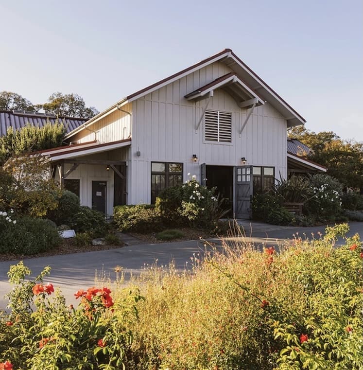 A charming white barn-style house with a red roof, nestled among lush greenery and blooming flowers in Healdsburg, California.