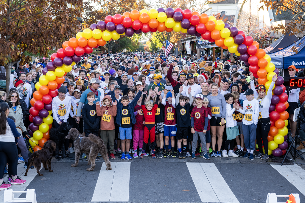 A large crowd of people, including children in superhero costumes, gathers under a colorful balloon arch for a race in Healdsburg, California.