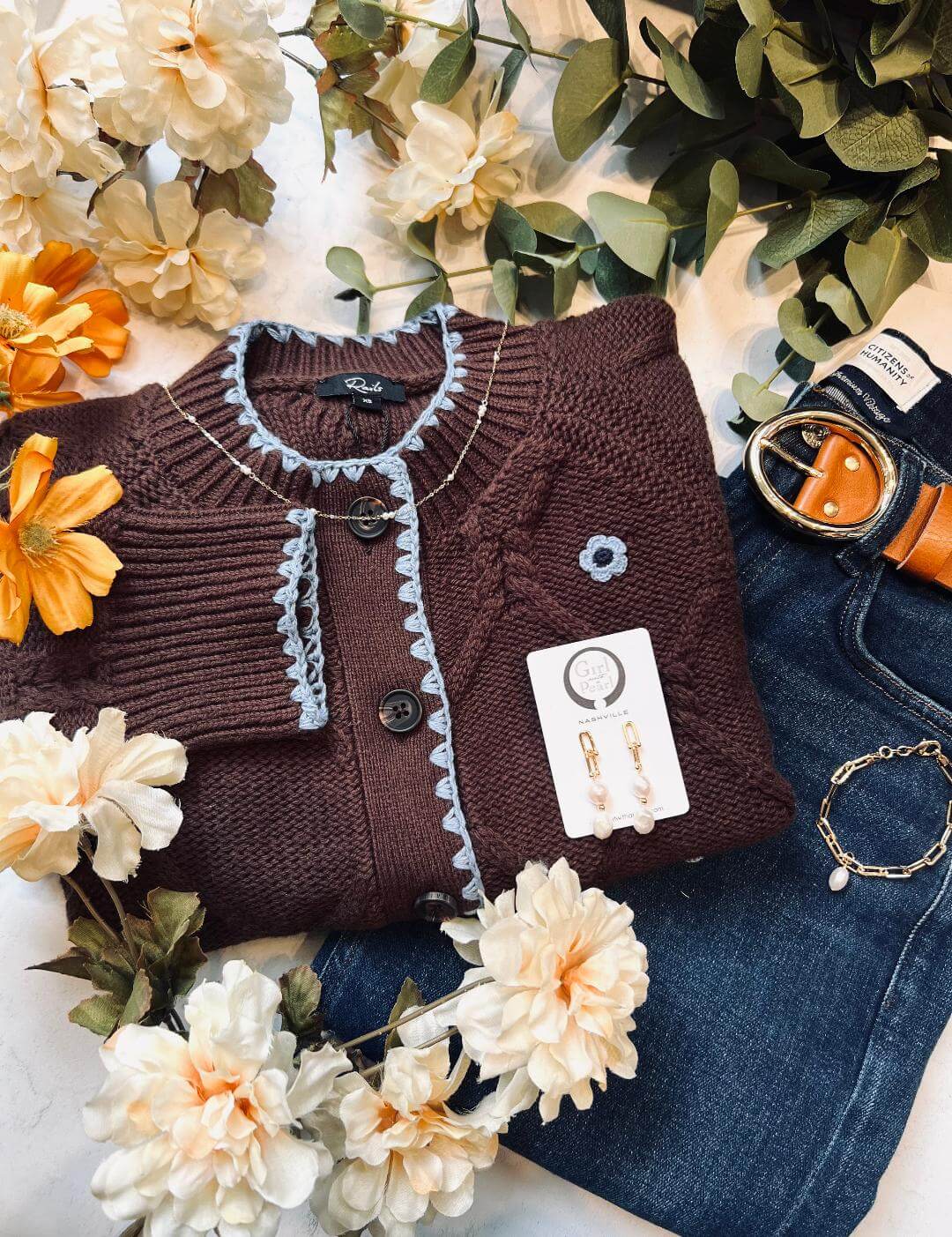 A stylish brown cable knit sweater with blue trim, adorned with white flowers and paired with dark blue jeans, a gold belt, and pearl earrings in Healdsburg, California.