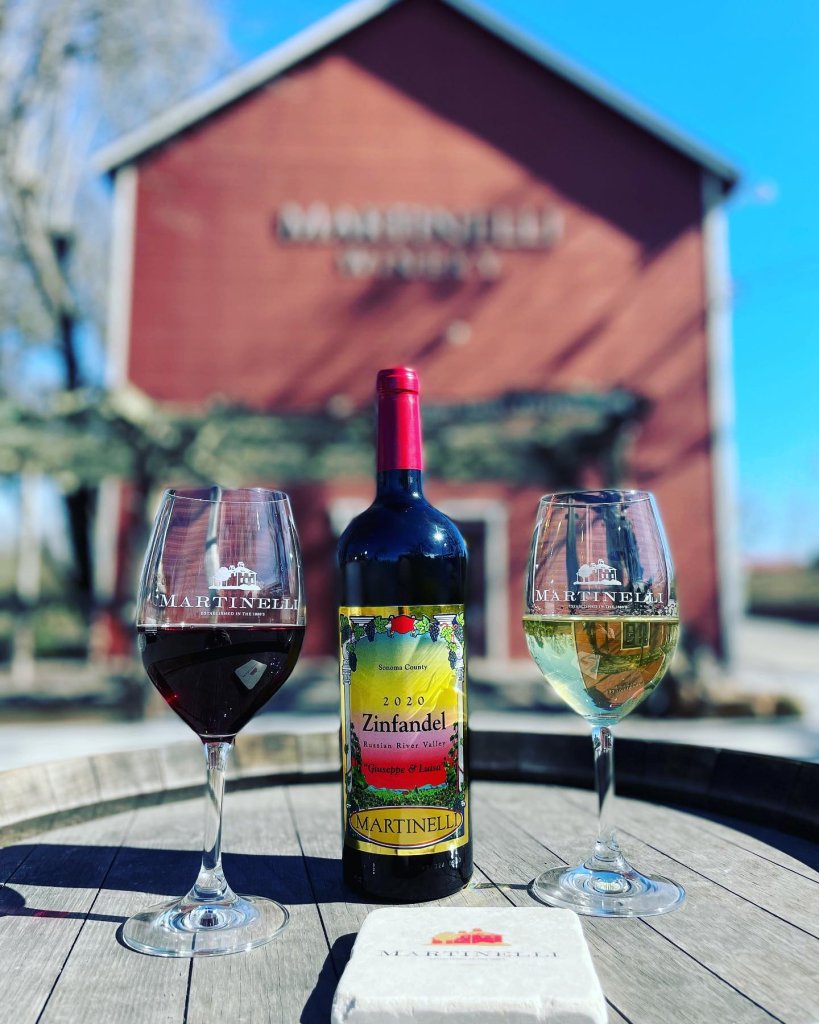 Two wine glasses and a bottle of 2020 Zinfandel sit on a wooden barrel in front of a red barn in Healdsburg, California.