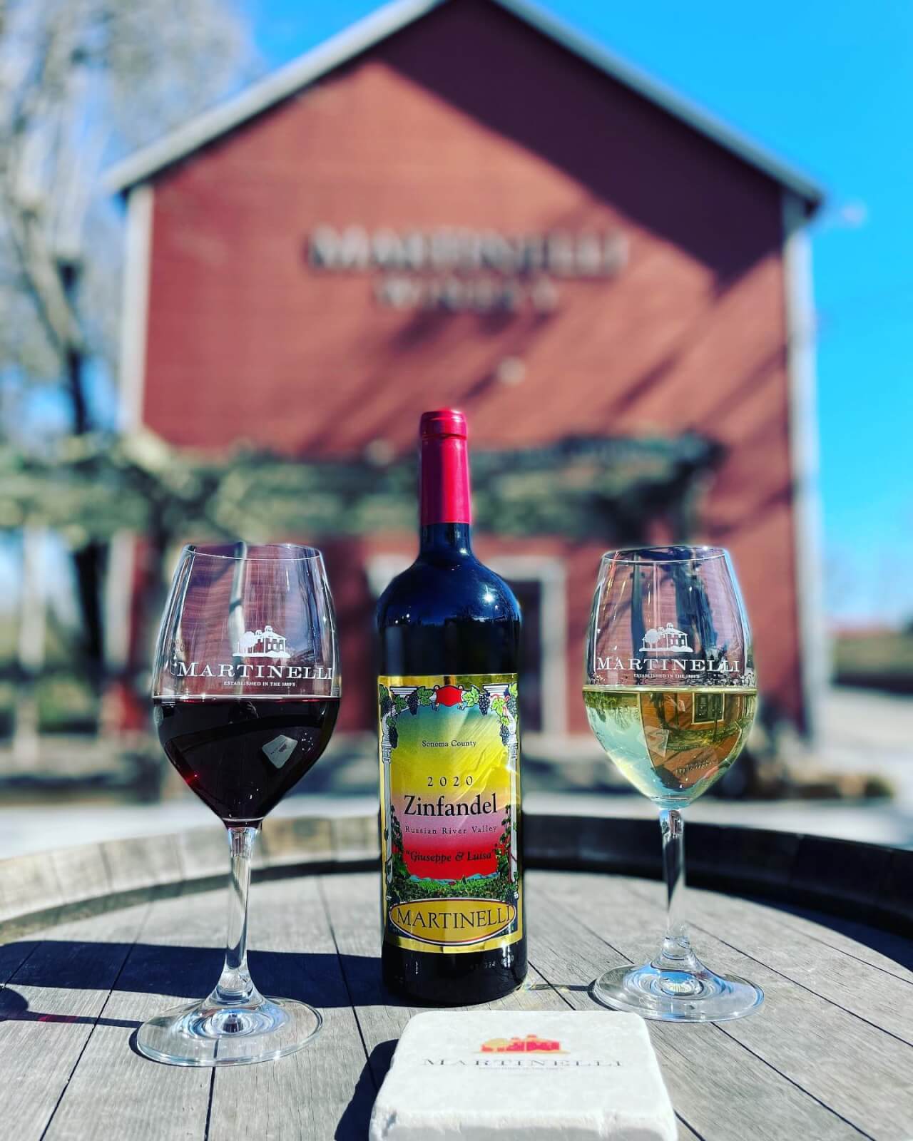 Martinelli Winery Two wine glasses and a bottle of 2020 Zinfandel sit on a wooden barrel in front of a red barn in Healdsburg, California.