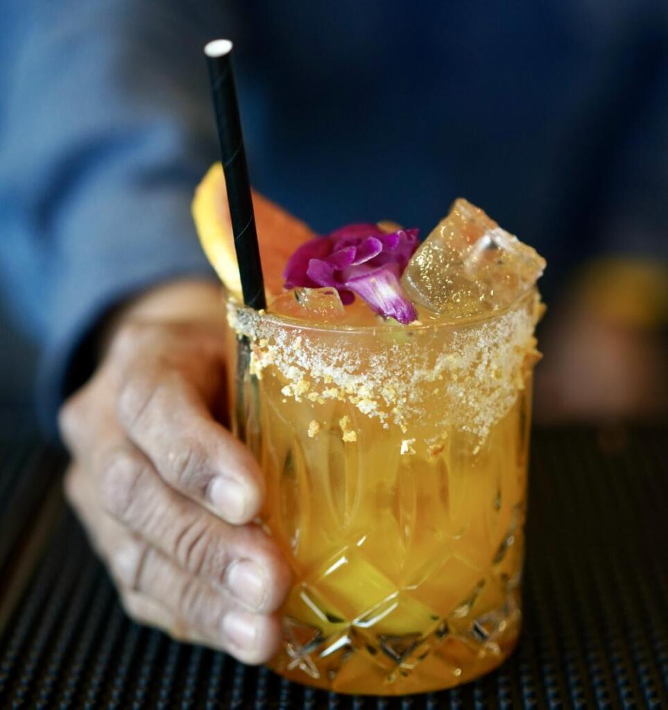 Mocktail Trail in Healdsburg A hand holds a glass of orange cocktail with ice, garnished with a flower and straw in Healdsburg, California.