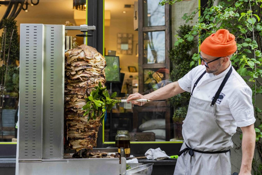 Chef in apron and beanie garnishes vertical rotisserie meat with herbs outside restaurant in Healdsburg, California.