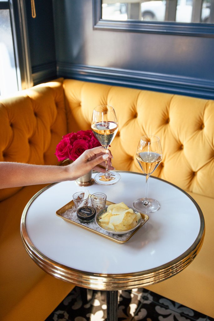 A woman in a yellow booth holds two glasses of white wine over a tray of caviar and chips in Healdsburg, California.