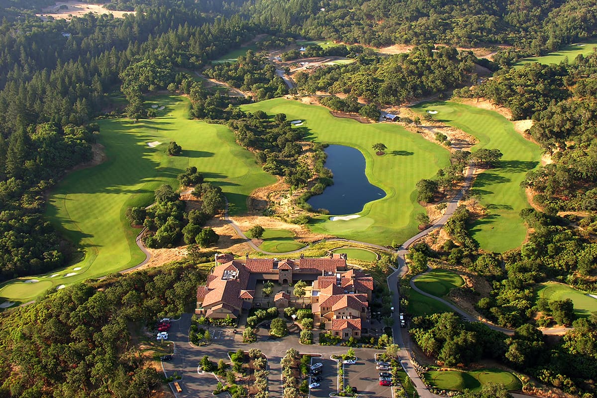 Stay & Play Special at The Ruse Aerial view of a sprawling golf resort with lush greens, a central lake, and a large Mediterranean-style clubhouse in Healdsburg, California.