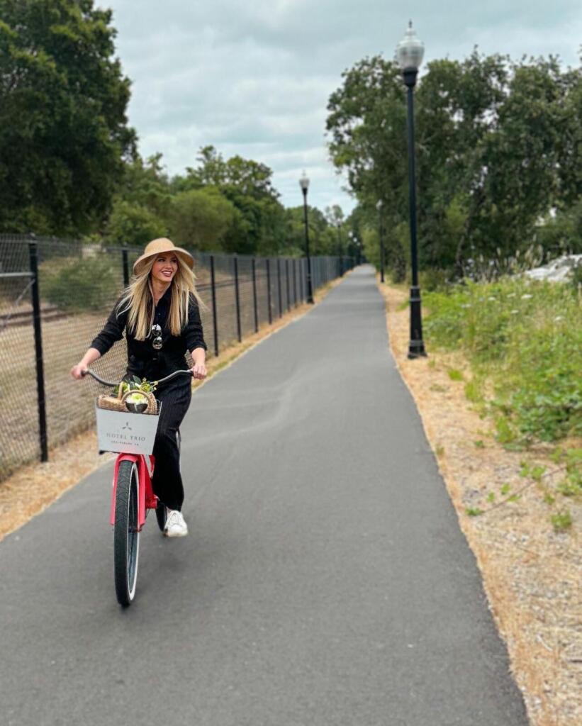 Discover the Best Bike Trails in Healdsburg A woman with long blonde hair rides a red bicycle down a paved path lined with trees in Healdsburg, California.