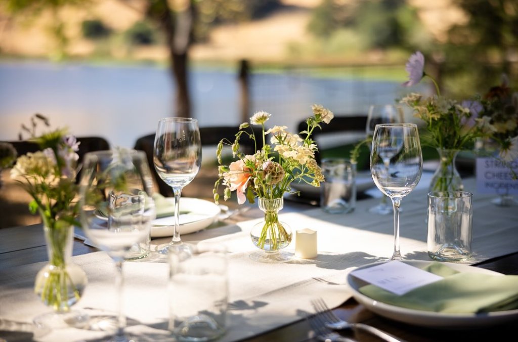 A beautifully set outdoor table with floral centerpieces and wine glasses overlooking a serene lake in Healdsburg, California.