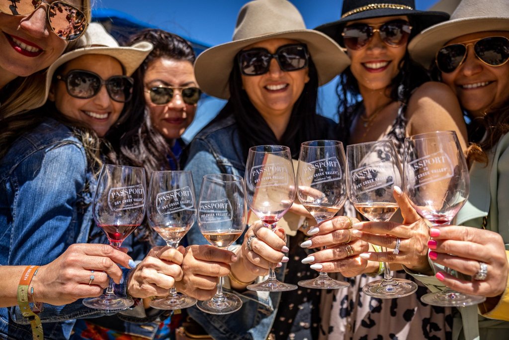 A group of six women, all wearing sunglasses and hats, cheerfully clink wine glasses together outdoors in Healdsburg, California.