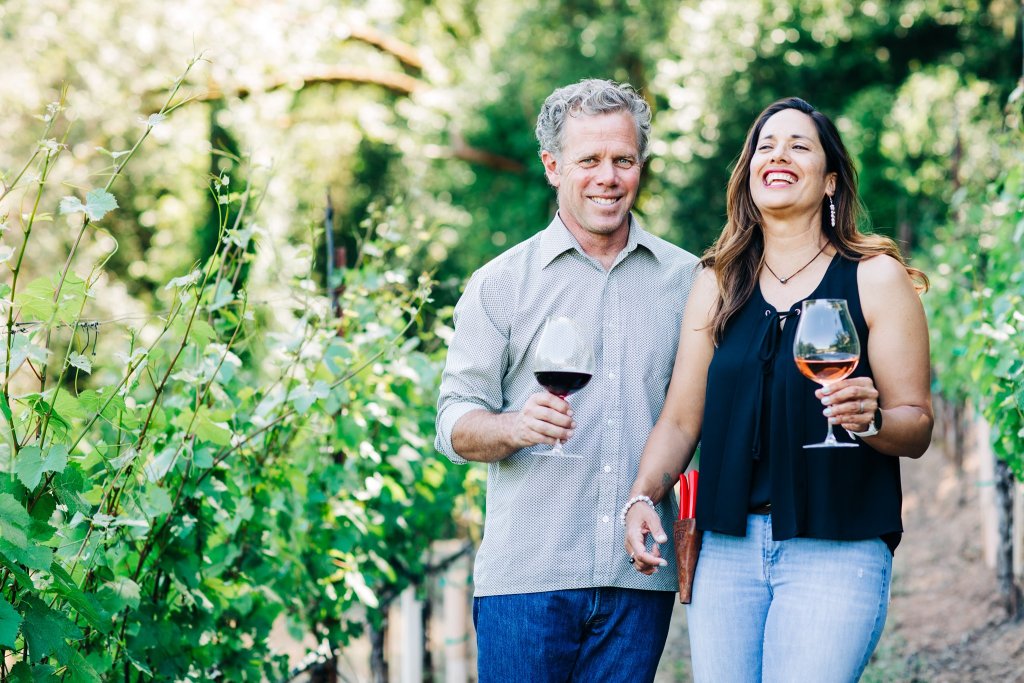 A man and woman smile while holding wine glasses in a lush vineyard in Healdsburg, California.