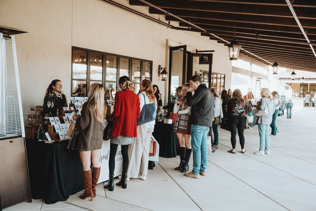 A group of people are gathered around tables at an outdoor event, browsing and purchasing items in Healdsburg, California.