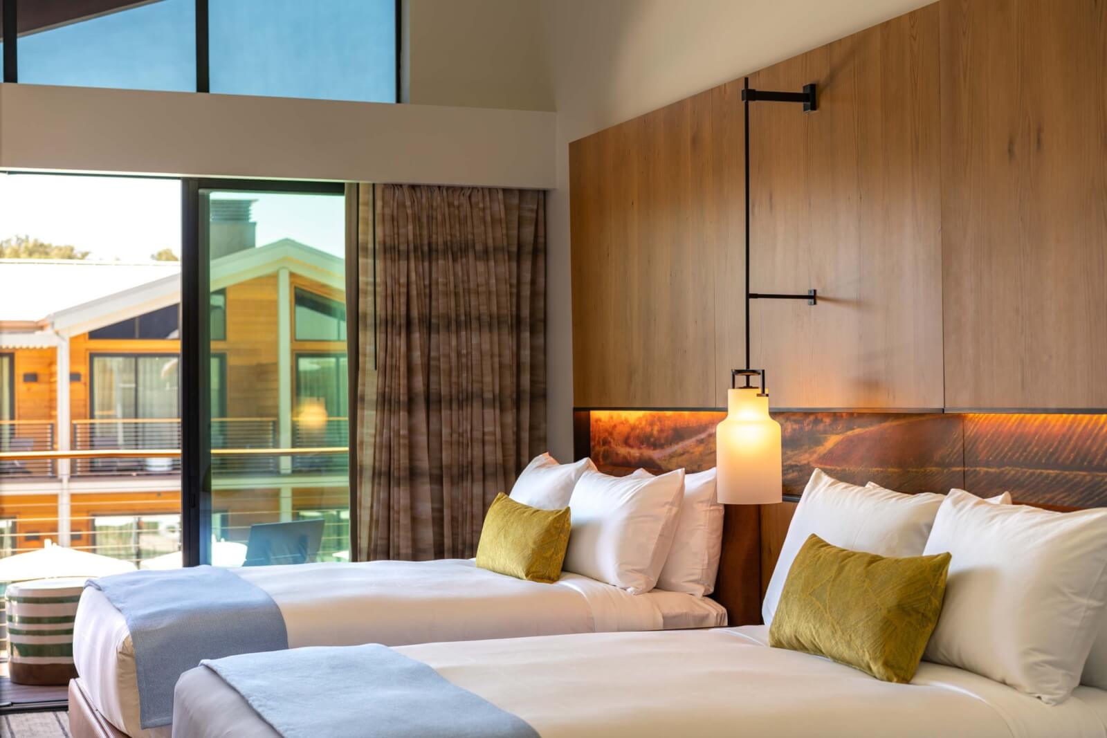 Appellation Healdsburg Modern hotel room with two beds, wooden headboard, and floor-to-ceiling windows overlooking a balcony and buildings in Healdsburg, California.