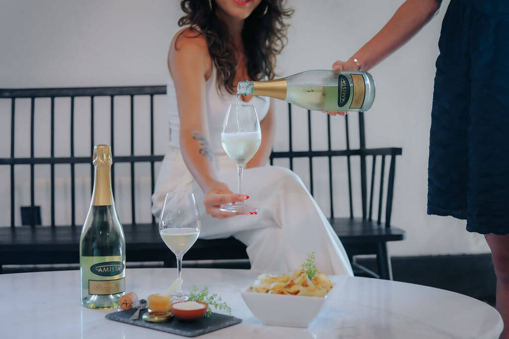 A woman in a white dress enjoys sparkling wine with a friend while snacking on chips and dips in Healdsburg, California.