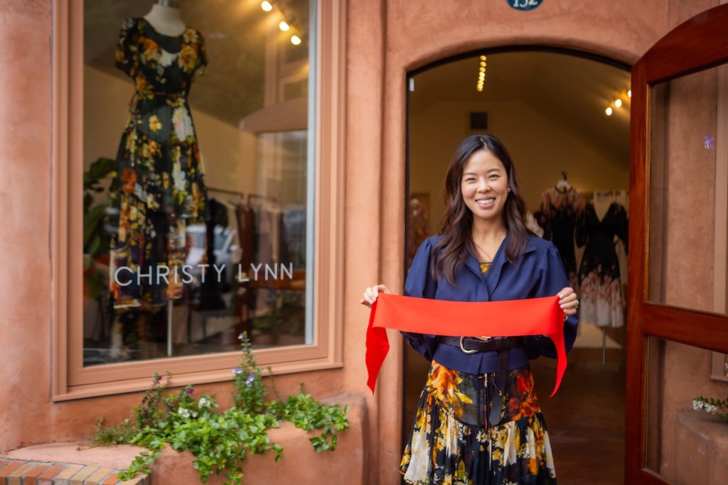 A woman stands in front of a clothing store holding a red ribbon, smiling in Healdsburg, California.