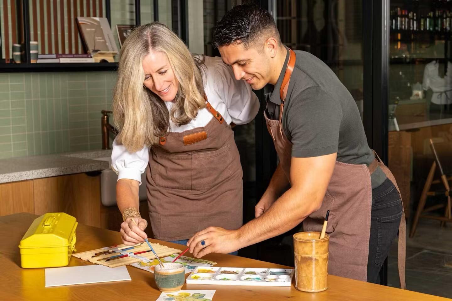 Two people in aprons paint together at a wooden table with art supplies in Healdsburg, California.