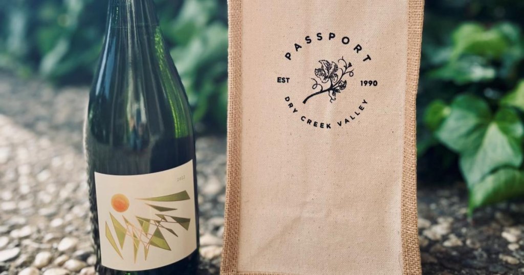 A green wine bottle with a geometric label stands next to a burlap bag with a floral logo in Healdsburg, California.