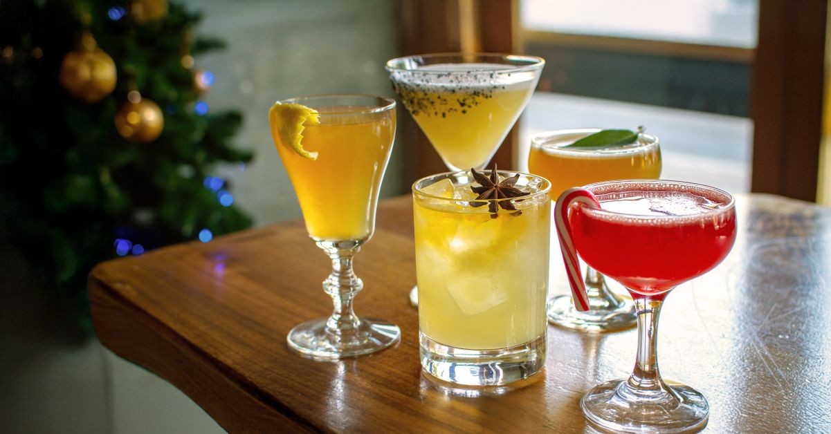 12 días de cócteles navideños en Spoonbar Five festive cocktails with fruit garnishes and spices on a wooden table near a Christmas tree in Healdsburg, California.
