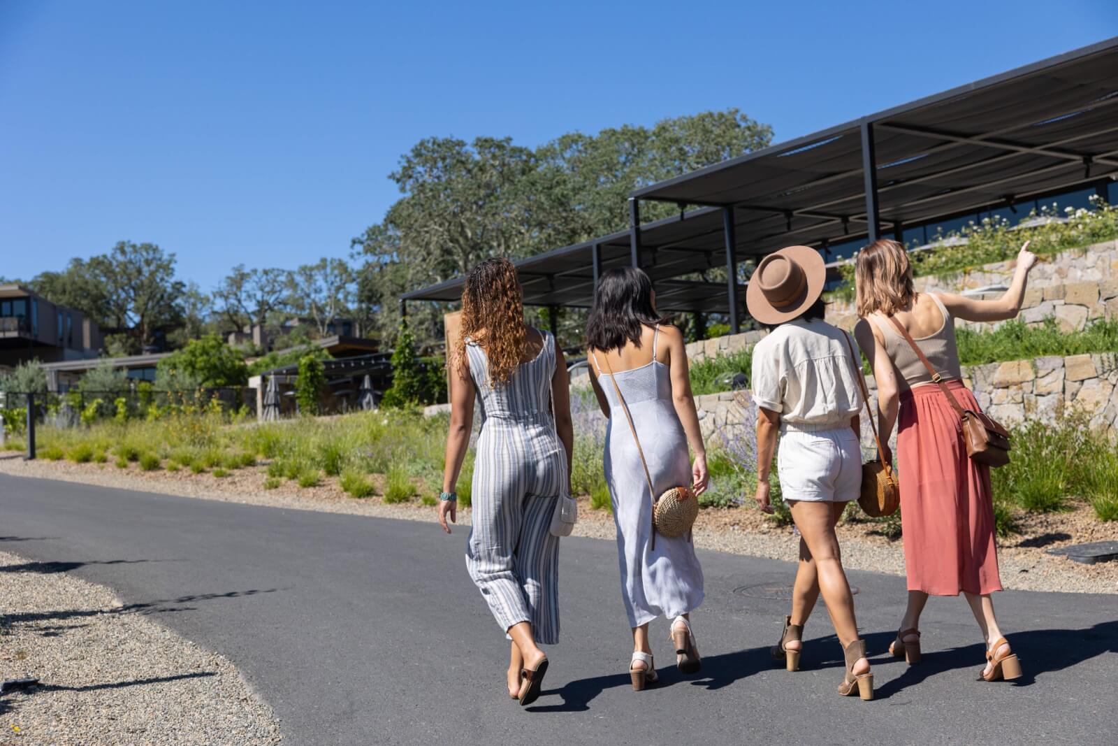 Four women in summer dresses and hats walk along a paved path in a sunny, green area in Healdsburg, California.