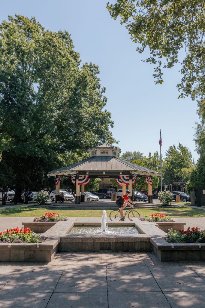 Prensa y medios A cyclist rides past a gazebo adorned with patriotic bunting in a park with a fountain and flower beds in Healdsburg, California.