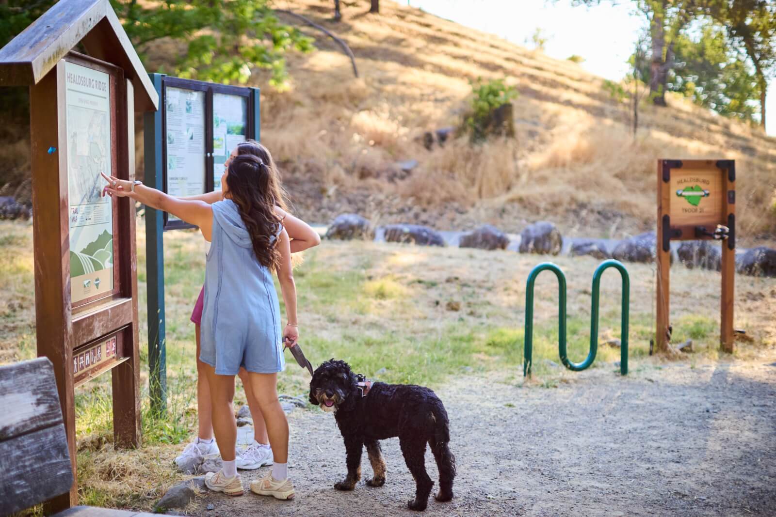 Healdsburg Blog Two women and a black dog stand near a wooden signpost with maps in a grassy outdoor area in Healdsburg, California.