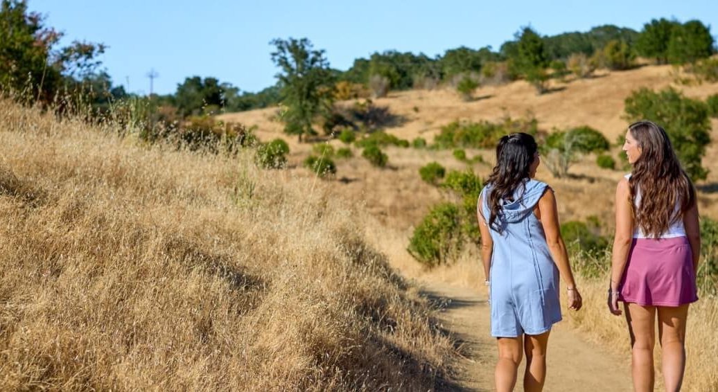 Two women in summer dresses walk along a dirt path through a dry, grassy hillside in Healdsburg, California.