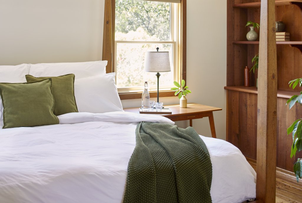 A serene bedroom with a white bed, green throw, wooden furniture, and a window overlooking greenery in Healdsburg, California.