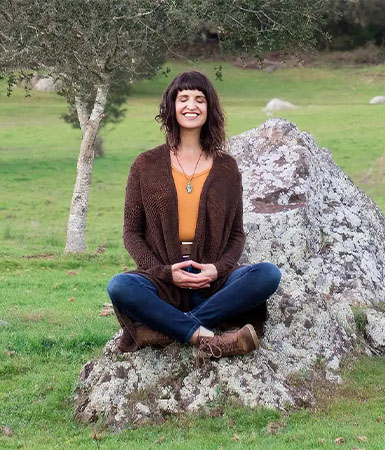 Hogar Woman meditating cross-legged on a rock in a grassy field with trees in Healdsburg, California.