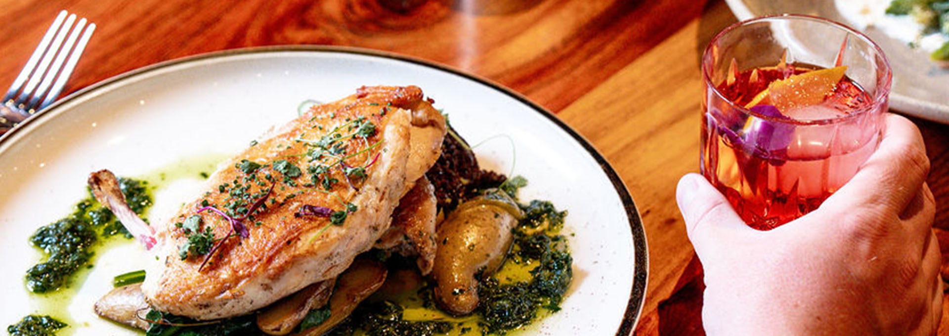 A hand holds a colorful cocktail next to a plate of roasted chicken with green sauce and vegetables in Healdsburg, California.