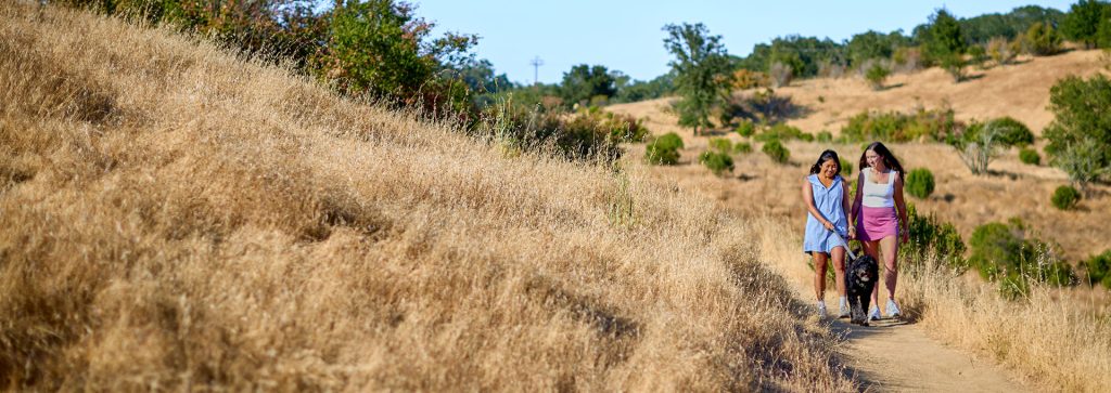 Two women and a dog walk along a dirt path through a dry, grassy hillside in Healdsburg, California.
