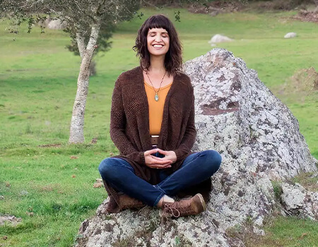 Woman sits cross-legged on a rock in a grassy field, smiling and meditating in Healdsburg, California.