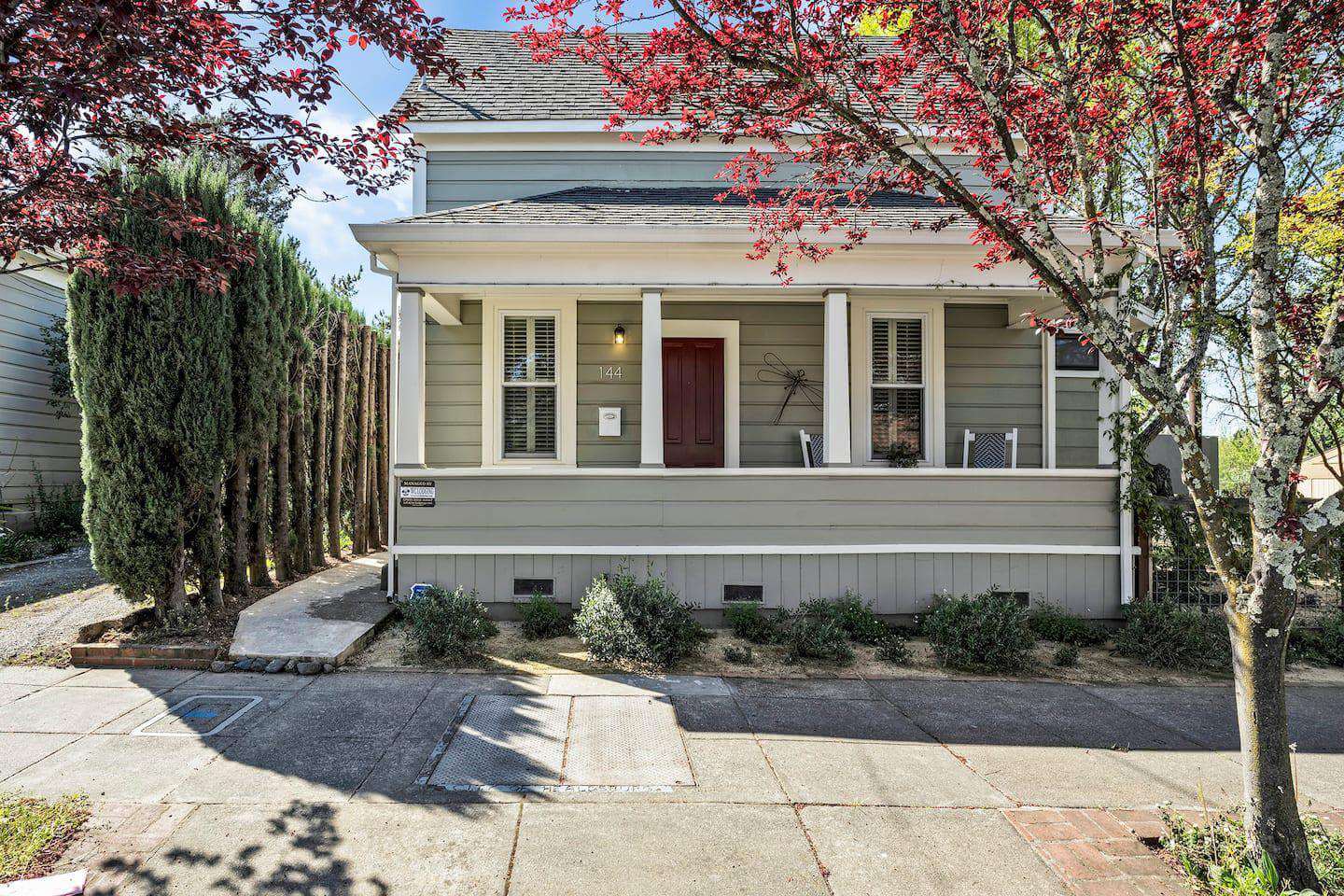 Carma Cottage A charming two-story house with a red door and white trim, nestled among trees and shrubs in Healdsburg, California.